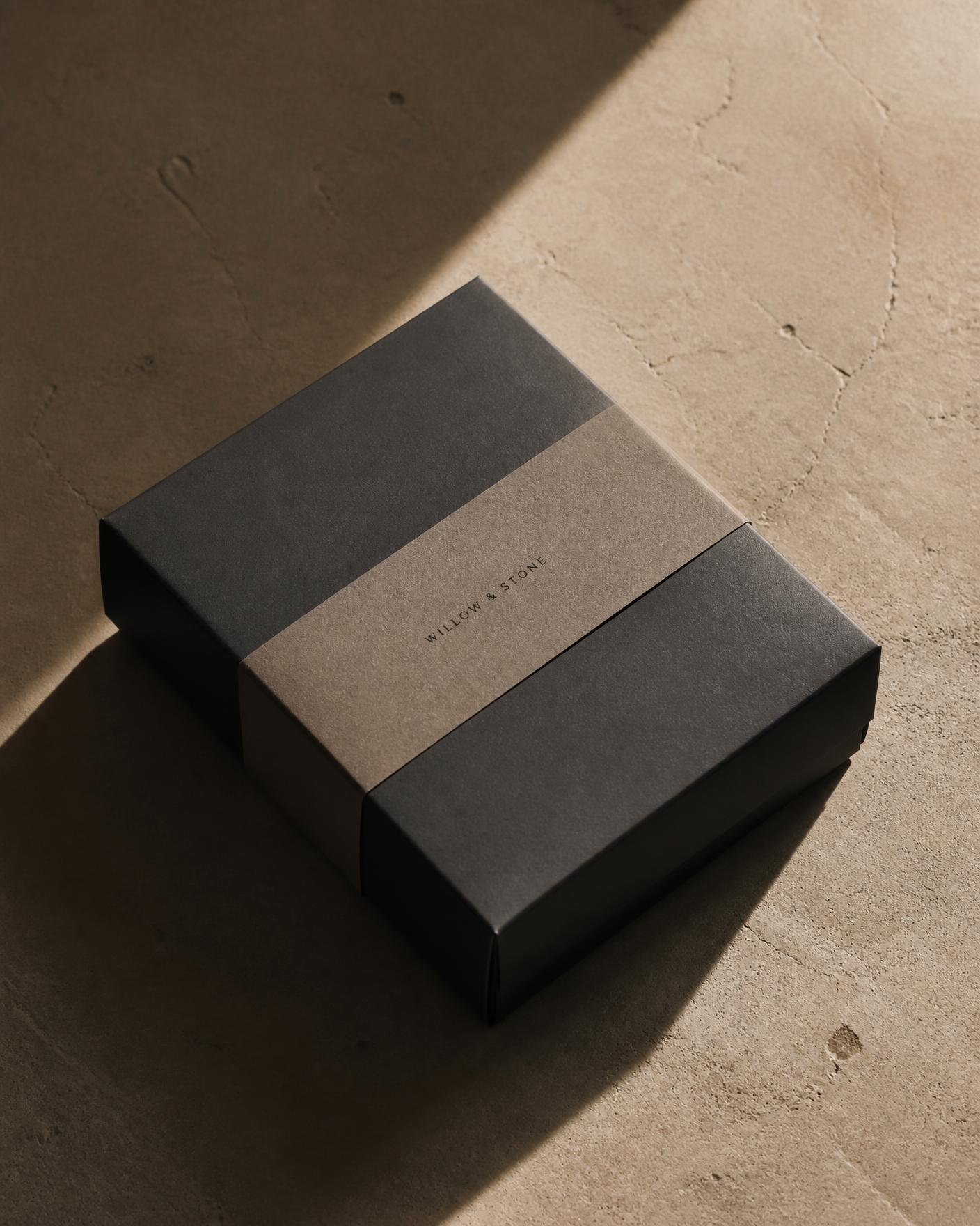 A matte charcoal gift box with a plain taupe band on a warm stone surface